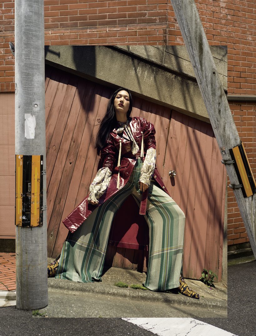 Person in a dark red leather coat and plaid trousers against a brick wall